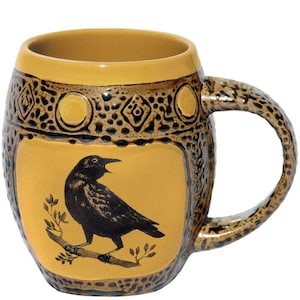 May include: A barrel-shaped ceramic mug featuring a mustard yellow glaze with a black illustration of a bird perched on a branch. The mug has a textured, dark brown speckled band around the top rim with geometric diamond and circle patterns.