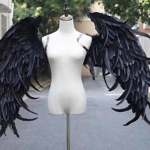 May include: Black feather angel wings displayed on a white mannequin. The wings are large and detailed, with a layered design. The mannequin is on a metal stand, set against a blurred outdoor background.