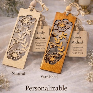 Può includere: Due segnalibri in legno, uno naturale e uno verniciato, con intagli floreali ed etichette allegate. Le etichette recitano "Emily & Michael, Engagement Keepsake, 06.15.2024." Il testo "Personalizzabile" è sotto.