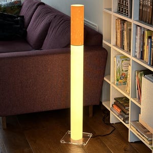May include: A tall, cylindrical floor lamp shaped like a cigarette, featuring a white illuminated base and an orange filter section at the top. The lamp stands on a clear, square acrylic base and emits a warm, soft glow from within the structure.