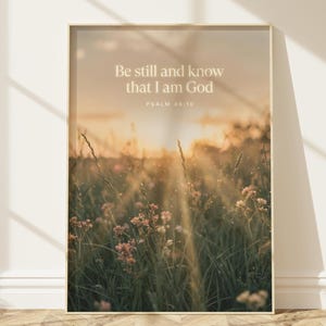 Op de afbeelding: Een rechthoekige wandkunstprint met een zonsonderganglandschap van een grasveld met wilde bloemen. De tekst "Be still and know that I am God" en "PSALM 46:10" is in het wit in het midden gedrukt tegen de gouden luchtachtergrond.