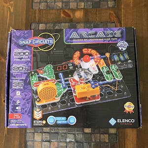 May include: A Snap Circuits Arcade electronics exploration kit box featuring a black background with purple accents. The box displays a circuit board with colourful components, a speaker, a fan, and a digital display. Text includes "ARCADE," "GAMES OF LEARNING AND FUN," and "OVER 200 EXCITING PROJECTS."