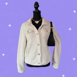 May include: A cream-colored sherpa jacket with a collared neckline, long sleeves, and a button-down front featuring metallic buttons. The jacket includes two chest pockets with flaps and is displayed on a black mannequin wearing a gold heart necklace.