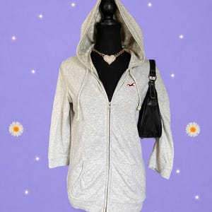 May include: A light gray zip-up hoodie with three-quarter length sleeves, a hood, and a small red bird logo on the chest. The hoodie is displayed on a mannequin wearing a gold-tone chain necklace with a heart pendant and a black shoulder bag.