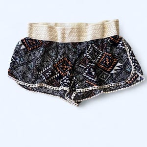 May include: A pair of patterned shorts with a cream-colored waistband and white pom-pom trim. The shorts feature a geometric design in black, white, and shades of orange and teal. The shorts are made of a lightweight fabric.