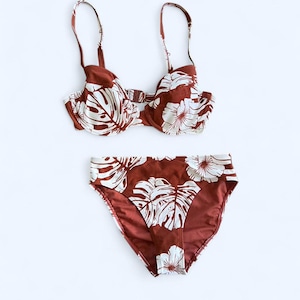May include: A two-piece swimsuit set with a brown and white floral design. The top is a bra-style with adjustable straps, and the bottoms are high-waisted. The pattern features large white leaves and flowers on a brown backdrop.