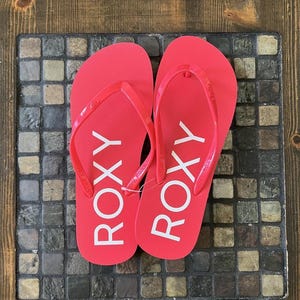 Peut inclure: Une paire de tongs rouge corail vif avec le nom de marque ROXY imprimé en grandes lettres capitales blanches sur la semelle. Les brides de type entre-doigts sont faites d'un matériau rouge corail assorti et incluent un petit logo en relief.