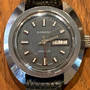 Könnte beinhalten: Eine Vintage Radiant Uhr mit dunkelgrauem Zifferblatt und silberfarbenem Gehäuse. Die Uhr zeigt Tag und Datum an, mit einem schwarzen Lederarmband. Das Zifferblatt hat weiße rechteckige Stundenmarkierungen.