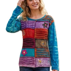 May include: A colourful patchwork long-sleeved top with floral details, in shades of blue, purple, red, and teal. The top is worn with blue jeans and sandals. A woven tote bag is held in the hand.