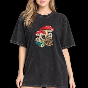 May include: A charcoal grey acid-wash t-shirt featuring a central graphic print of two red and white spotted mushrooms, a brown snail, and small white flowers with green leaves. The garment has a crew neckline and short sleeves with a relaxed fit.