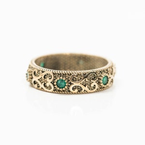 May include: A gold-coloured ring with an ornate design featuring scrollwork and small, round, emerald-green gemstones. The ring has a textured band and is set against a white background.