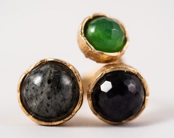 Può includere: Un anello d'oro con tre pietre: una pietra verde, una pietra nera e una pietra grigia.