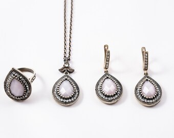 May include: A set of jewelry featuring a teardrop-shaped pendant necklace, earrings, and ring. Each piece has a clear, faceted stone set in a silver-toned metal frame with a decorative border of smaller stones.