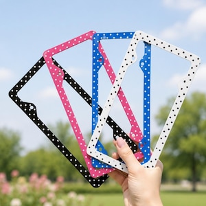 May include: Four license plate frames in black, pink, blue, and white, each with a polka dot pattern. The frames are held up against a blue sky and green background.