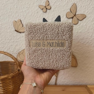 Peut inclure: Un objet carré en tissu sherpa beige texturé, doté d'une étiquette rectangulaire avec le texte noir Luise & Mathilde cousu au centre. Deux petites formes triangulaires sombres dépassent du bord supérieur de la matière douce et duveteuse.
