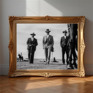 May include: Black and white framed print featuring three men in suits and hats walking across a field. The ornate gold frame adds a vintage aesthetic. A horse and rider are visible in the background.