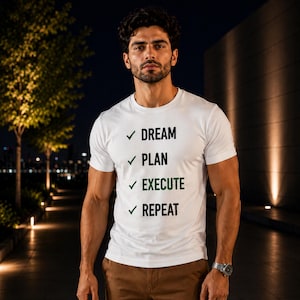May include: A white short-sleeved t-shirt featuring a list with green checkmarks next to the words DREAM, PLAN, EXECUTE, and REPEAT printed in black and green text on the chest. The shirt is paired with brown trousers and a silver wristwatch.