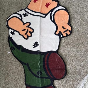 Könnte beinhalten: Ein ausgefallener Teppich in Form einer Zeichentrickfigur. Der Teppich zeigt eine Figur mit weißem Oberteil, grüner Hose und braunen Schuhen. Gesicht und Arme sind pfirsichfarben, mit schwarzen Umrissen und roten Details.