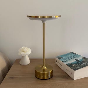 Peut inclure: Une lampe de chevet dorée avec un dessus et une base ronds, et une tige fine. Un vase en céramique blanc avec une fleur blanche et un livre sont sur la table de chevet en bois. Le livre est intitulé "Le Pacte de L'eau".