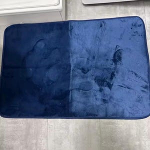 Op de afbeelding: Een rechthoekige, marineblauwe badmat. De mat heeft een zachte, fluweelzachte textuur en lijkt gedeeltelijk gereinigd te zijn, met een zichtbaar contrast tussen de schone en vuile delen. De mat is ontworpen voor gebruik in de badkamer.
