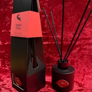 Könnte beinhalten: Ein schwarzes Duft-Diffusor-Set mit passender Box. Die Box hat ein rotes Etikett mit dem Text "Opelia's Candy" und einem Kaninchen-Logo. Die Diffusorflasche und die Box haben ein rotes Siegel. Der Diffusor hat schwarze Stäbchen.