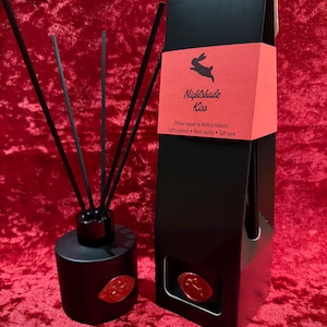 May include: A black diffuser set with reeds and a matching box, both featuring a red label with a rabbit silhouette and the text "Nightshade Kiss." The set includes a round bottle with a red seal.