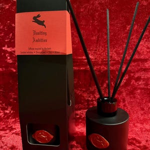 Könnte beinhalten: Schwarzes Reed-Diffusor-Set mit passender Box. Die Box hat ein rotes Etikett mit einem Hasenlogo und dem Text "Vaunting Ambition". Der Diffusor ist schwarz mit schwarzen Stäbchen und einem roten Wachssiegel.