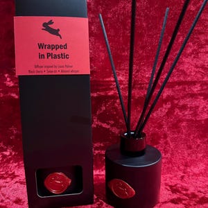 Op de afbeelding: Zwarte diffuser met stokjes en een bijpassende doos. De doos heeft een rood label met de tekst "Wrapped in Plastic" en een konijnlogo. De diffuser is geïnspireerd door Laura Palmer en heeft geuren van zwarte kers, sneeuwlucht en amandelwhisper.