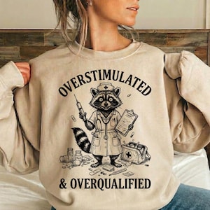 以下が含まれることがあります： 看護師の格好をしたアライグマのグラフィックが描かれたベージュのスウェットシャツ。注射器とチャートを持っています。「OVERSTIMULATED & OVERQUALIFIED」の文字がアライグマのイラストの上と下にプリントされています。医療用品が散らばっています。