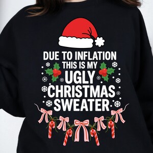 May include: A black long-sleeve sweatshirt featuring a graphic print of a red Santa hat above the text "DUE TO INFLATION THIS IS MY UGLY CHRISTMAS SWEATER" with holly leaves, berries, white snowflakes, pink bows, and red and white candy canes.