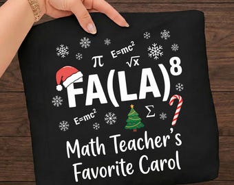 Op de afbeelding: Een zwart t-shirt met witte tekst FA(LA)^8 en Math Teacher's Favorite Carol. Het ontwerp bevat wiskundige symbolen zoals pi, vierkantswortel, E=mc2 en sigma, naast een rode kerstmuts, een groene kerstboom en een zuurstok.