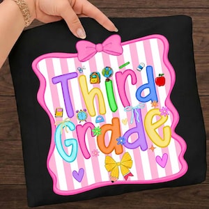 Könnte beinhalten: Ein schwarzes T-Shirt mit einem Aufdruck, der den Text "Third Grade" in bunten Buchstaben zeigt. Das Design umfasst einen pink-weiß gestreiften Hintergrund, eine pinke Schleife oben, eine gelbe Schleife unten und verschiedene Schulmotive.