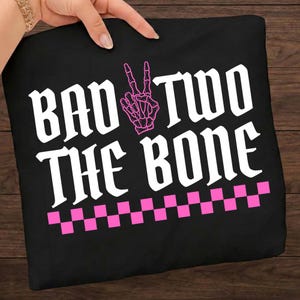 May include: Black t-shirt with the phrase "BAD TWO THE BONE" in white gothic font. A pink skeleton hand making a peace sign is above the word "TWO". A pink and white checkered pattern is at the bottom of the design.