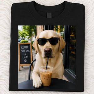 May include: A black t-shirt featuring a graphic of a yellow Labrador dog wearing black sunglasses and drinking an iced coffee from a clear plastic cup with a straw. In the background, a chalkboard sign displays the text Coffee Good Days & Labs.