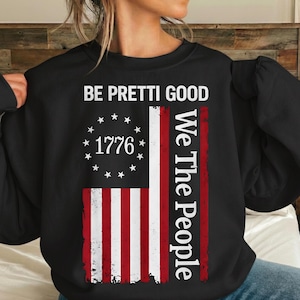 Puede incluir: Sudadera negra con un diseño desgastado de la bandera estadounidense. La bandera incluye rayas rojas y blancas, un círculo de estrellas y el año "1776". El texto en la sudadera dice "BE PRETTI GOOD" y "We The People."
