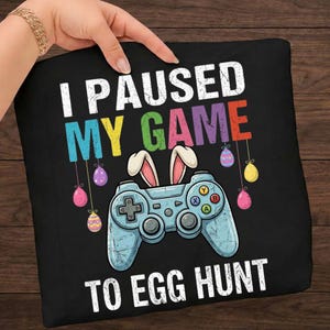 Könnte beinhalten: Schwarzes Kissen mit dem Text "I PAUSED MY GAME TO EGG HUNT" in Weiß, Blau, Gelb, Grün und Rot. Ein hellblauer Gamecontroller mit Hasenohren und Ostereiern.
