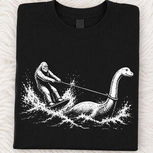 May include: A black crew neck t-shirt featuring a white graphic illustration of a hairy creature resembling Bigfoot water skiing while being pulled by a long-necked aquatic creature, both surrounded by detailed white water splashes and waves.