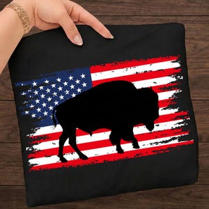 May include: Black cushion featuring a distressed American flag design with a silhouette of a bison. The flag has red and white stripes and a blue field with white stars. The bison is a solid black colour.