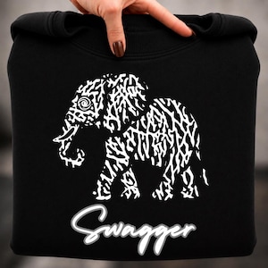 May include: A black sweatshirt featuring a white graphic of an elephant composed of intricate, abstract line patterns. Below the elephant, the word "Swagger" is printed in a white, cursive script font with a subtle grey outline on the fabric.