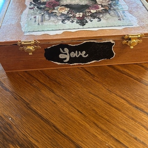 May include: Wooden box with a decorative lid featuring a heart design with floral accents in pink, white, and black. The box has gold-colored clasps and the word "Love" written on a black label.