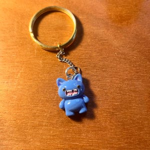 May include: A blue cartoon cat keyring with a gold-coloured ring. The cat has a wide, toothy grin and small arms and legs. The keyring is attached to a small silver chain.