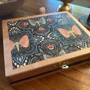 May include: A wooden box with a decorative lid featuring a butterfly and floral pattern in shades of pink, blue, and gold. The box has a brass clasp and a light brown wooden frame. The box is sitting on a wooden surface.