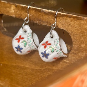 May include: A pair of dangle earrings featuring miniature white ceramic teacup charms. Each teacup is decorated with small red and blue floral patterns, green leaves, and gold-toned accents. The charms hang from silver-toned metal fishhook wires.
