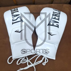 Puede incluir: Par de guantes de boxeo blancos Everlast con letras y logotipos negros. Los guantes tienen cordones blancos y están hechos de un material liso. Los guantes están sobre una superficie marrón.