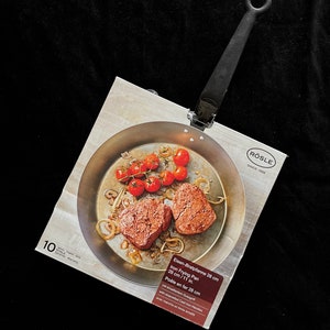 May include: An iron frying pan with a long handle, displayed on its packaging. The pan contains cooked food, including tomatoes and meat. The packaging reads "Iron Frying Pan 28 cm / 11 in." and the brand name "RÖSLE".