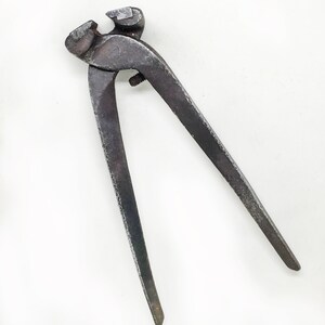 May include: A pair of vintage metal pliers with a worn, dark gray finish. The pliers have a simple design with a single rivet connecting the handles.