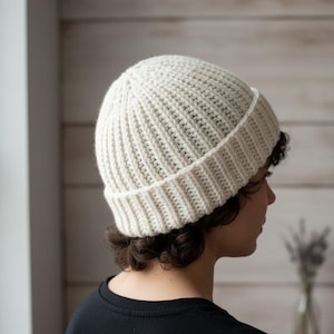 May include: Cream-coloured, ribbed knit beanie hat. The hat is made of a thick, textured yarn and features a folded brim. The hat is worn by a person with dark hair and a black top. The background is a light-coloured wooden wall.