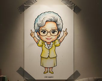 May include: Illustration of a woman in a yellow suit with glasses, arms raised in a peace sign. The artwork is taped to a wall with gray tape. The text "CORY AQUINO" is at the bottom.