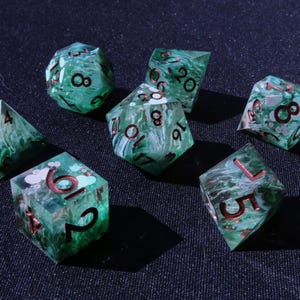 May include: Set of seven polyhedral dice in shades of green with white marbling and red numbering. The dice are designed for tabletop role-playing games. The dice include a d4, d6, d8, d10, d12, d20, and a percentile die.