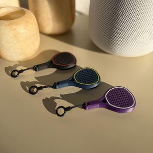 Könnte beinhalten: Drei Miniatur-Tennis-Schlüsselanhänger in Schwarz, Blau und Lila, jeweils mit kontrastfarbener Umrandung. Die Schläger haben eine strukturierte Oberfläche und sind an einer schwarzen Schlaufe befestigt. Die Schlüsselanhänger werden auf einer hellbeigen Oberfläche präsentiert.